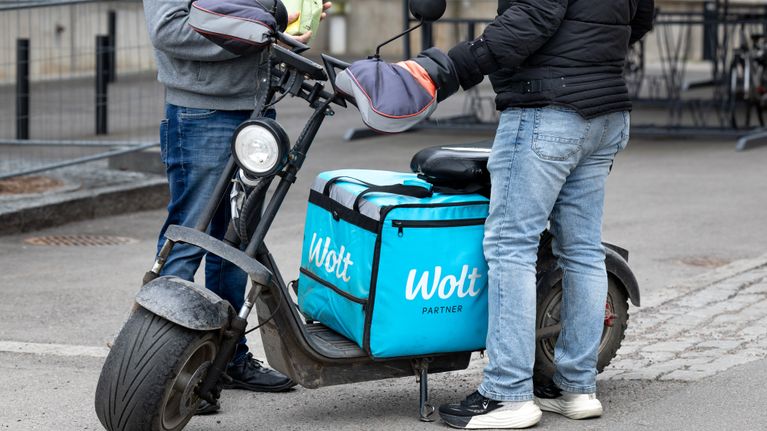 Two people stand by an electric scooter with a light-blue carrying case with the words Wolt Partner. Their heads are cropped out of the picture.