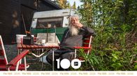 Man med grått skägg sitter i en stol framför en husvagn och röker en cigarrett. Yle100-grafik i nedre kanten.