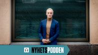 En man iklädd en blå kavaj och en beige tröja står vid en byggnad. Han lutar sig lätt mot väggen medan han håller händerna framför sig.