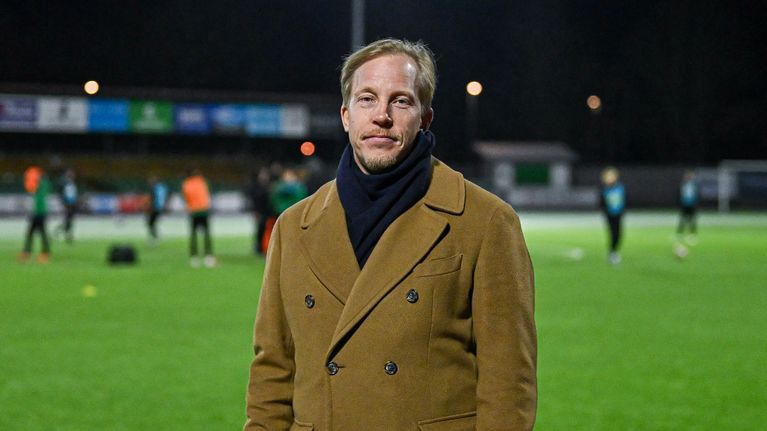 En man står på en fotbollsplan, klädd i en ljusbrun dubbelknäppt kappa och en mörk halsduk.