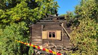 En nedbrunnen gammal timmerbyggnad avspärrad med polistejp, omgiven av grönska.