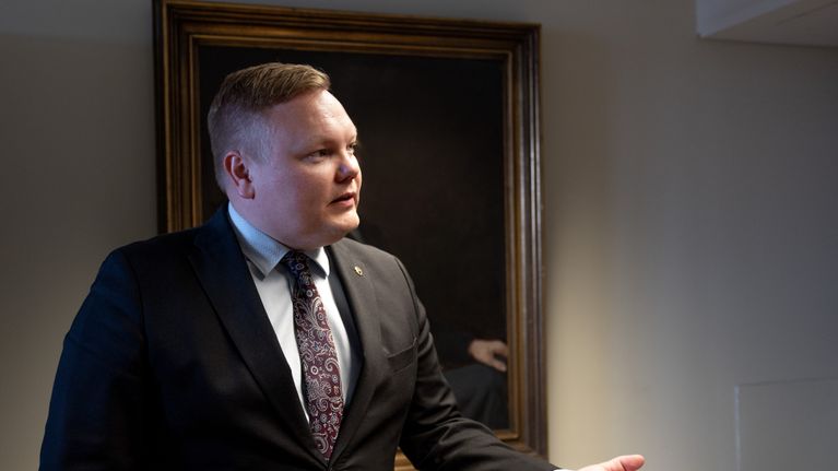 Puvuntakkiin pukeutunut mies puhuu sisätiloissa taulun edessä.