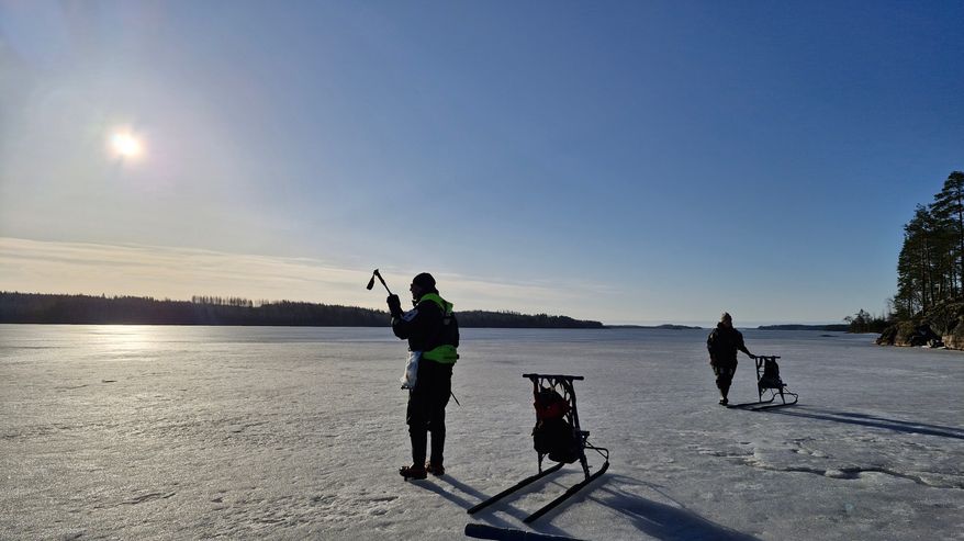 Kaksi henkilöä seisovat järven jäällä kuivapuku päällään. Vieressä on potkukelkat. Aurinko paistaa.