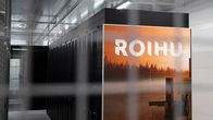 Roihu-supertietokoneen laitekaappi, jonka kylkeen on teipattu oranssi maisemakuva ja Roihu-teksti, näkyy metallisen verkkoaidan läpi.