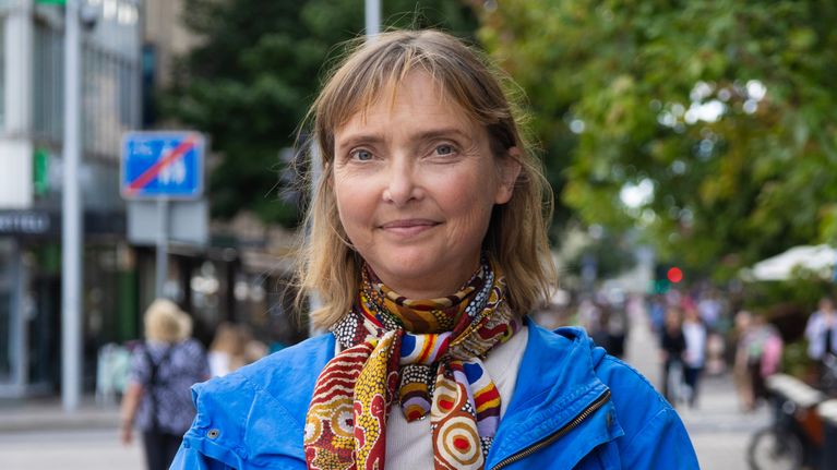 En kvinna med blå jacka och färgglad scarf står på en trottoar med butiker och träd i bakgrunden.