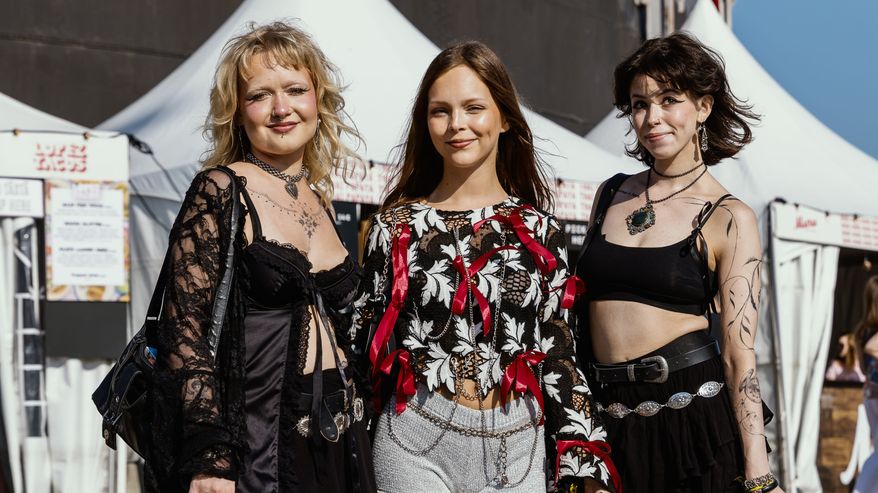 Kolme naista poseeraa ulkona festivaalialueella tyylikkäissä ja vaihtoehtoisissa asuissa, yhdistäen mustia ja värikkäitä vaatteita sekä pitsiä ja nahkaa. Taustalla näkyy valkoisia telttoja.