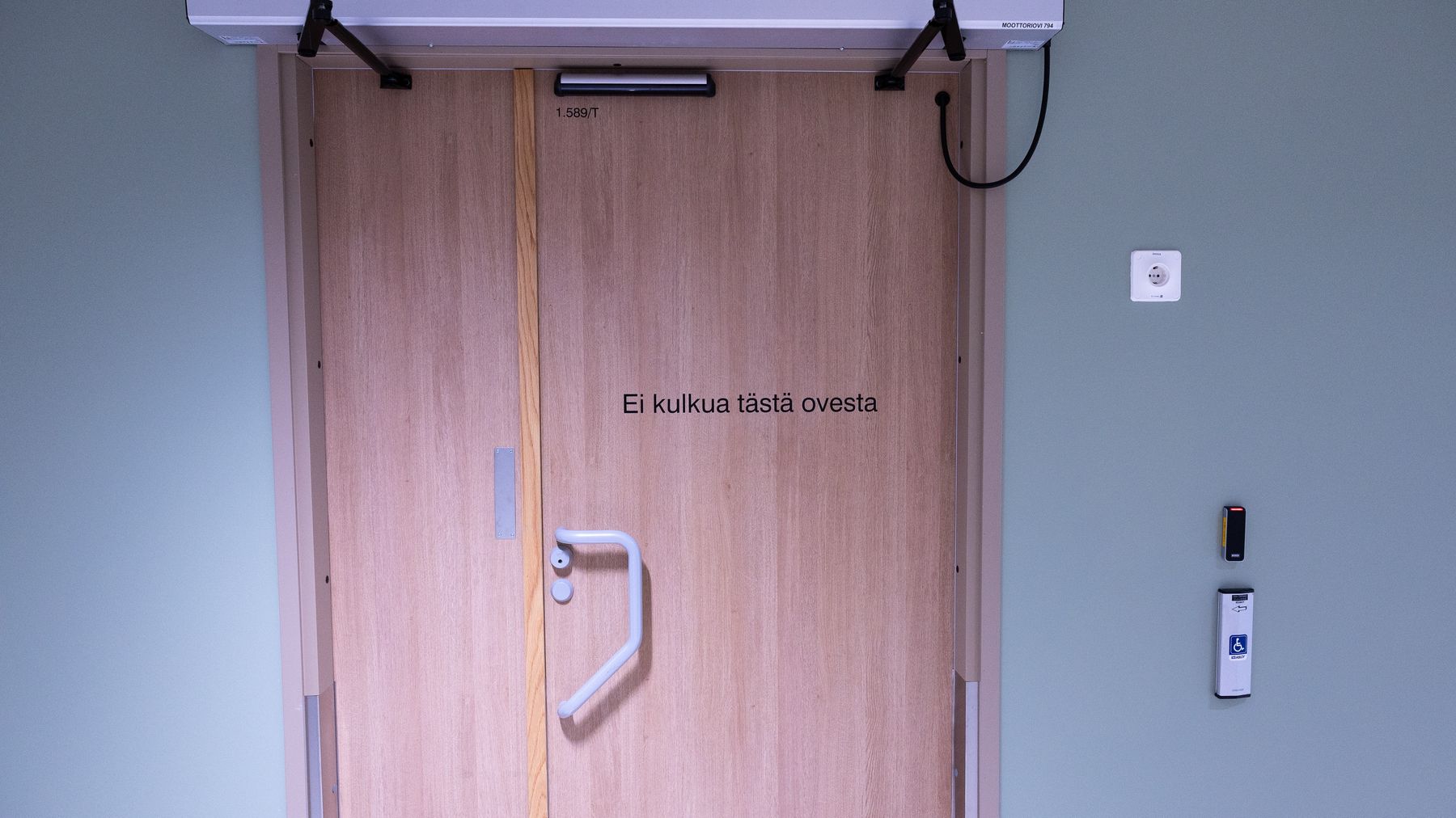 Suljettu puuovi, jonka yläpuolella on laite, ovessa lukee teksti "Ei kulkua tästä ovesta", seinässä vieressä sähköinen kulunvalvontalaite.