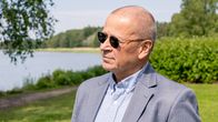 Henkilö pukeutunut harmaaseen pukuun ja aurinkolaseihin seisoo ulkona järvimaisemassa, taustalla vihreää kasvillisuutta.