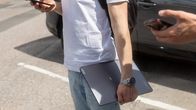 En person står på en parkeringsplats med telefonen i ena handeln och håller en hopslagen laptop i den andra. I kanten av bilden syns en annan hand med telefon. Båda är fotade så att huvudet inte syns på bilden.