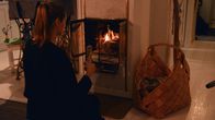 Merja Forsman sitter på knä framför kakelugnen och fyller på ved i brasan. 