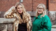 Två kvinnor står utomhus framför en tegelbyggnad; den ena bär pälsjacka, den andra grön jacka.