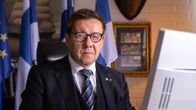 Tummaan pukuun pukeutunut Jari Andersson istumassa tietokoneen ääressä katse kameraan. Taustalla lippuja, tauluja ja täytetty hirven pää.