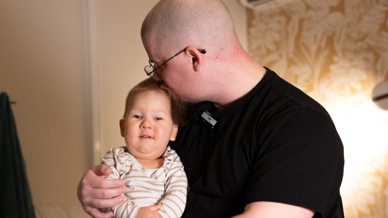 Mies istuu tuolilla pitäen sylissään lasta olohuoneessa.