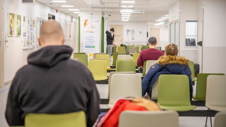 Three people sitting in a hospital waiting room, with a person standing in the background.