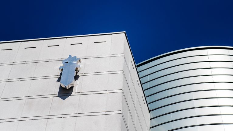 The top of white building with a square and a round section with a sword logo, seen against a clear blue sky. 