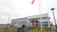 Kolme ihmistä seisoo Danfoss-nimisen rakennuksen edessä, lipputangossa punainen lippu, pihalla näkyy nurmialueita.