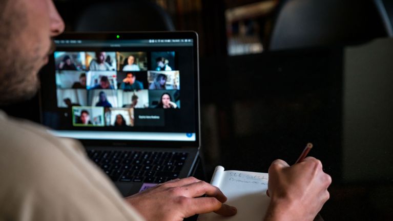 En ung man sitter framför en bärbar dator och antecknar i ett skrivblock. På datorn syns programmet zoom öppnat med flera videobilder av människor.
