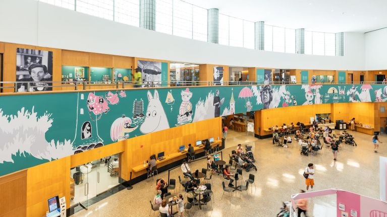 Muumi 80-juhlavuoden suurnäyttely Brooklyn Public Library -kirjastossa. Tilan seinää koristaa muumien ja Tove Janssonin kuvat, ihmisiä kävelee kirjastossa ja istuu pöytäryhmien ääressä.