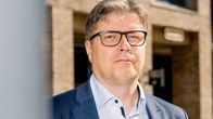 Mies seisoo ulkona tiilirakennuksen edessä, päällään sininen puku ja vaaleansininen kauluspaita. 
