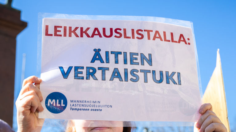 Nainen pitää kädessään kylttiä, jossa lukee "Leikkauslistalla: äitien vertaistuki".