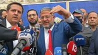 Group of men behind several media microphones, one man in a blue suit jacket and red necktie is gesturing with his hand as he speaks.