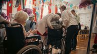 Four elderly people seen from behind, seated in wheelchairs with three women standing and helping them in a decorated common room.
