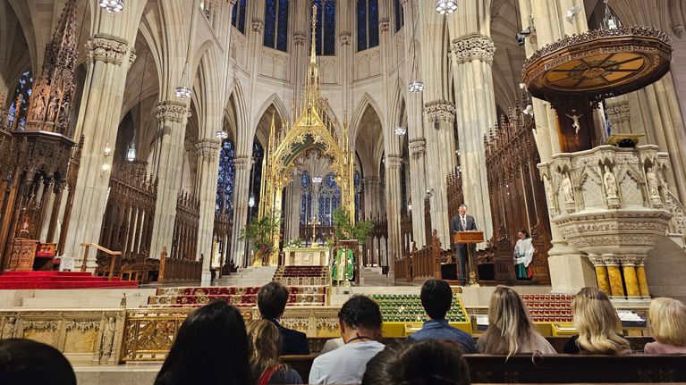 En gudstjänst pågår i en nygotisk kyrka med förgylld konstruktion ovanför högaltaret. I de främsta bänkraderna sitter gudstjänstbesökare.