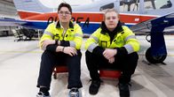 Kaksi henkilöä istuu lentokonehallissa lentokoneen edessä, heillä on päällään keltaiset huomiotakit.