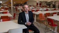 Mies istuu tyhjässä lukiossa, taustalla näkyy portaikko ja tyhjiä pöytiä ja tuoleja.