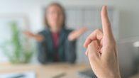 A closeup of an outstretched index finger in front of a blurred woman sitting in the background. 