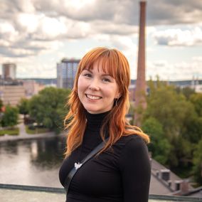 Nainen seisoo kattoterassilla ja hymyilee kameralle, taustalla on punatiilinen savupiippu, puita, rakennuksia ja Tammerkoski.
