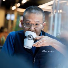 Man with short hair and eyeglasses in a blue shirt drinking from a white coffee mug, looking at the camera.