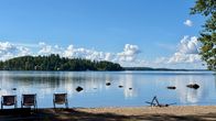 Hiekkaranta, neljä rantatuolia ja nuotiopaikka järven rannalla; taustalla metsäinen saari ja pilvinen taivas.