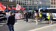 Mielenosoittajia punaisten lippujen ja banderollien kanssa kadulla poliisiauton edessä.