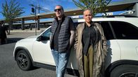 Två personer står lutade mot en vit bil på en parkeringsplats med träd och byggnader i bakgrunden.