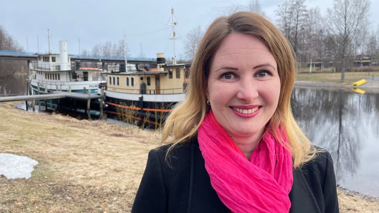 En kvinna med blont hår står vid en å och ler medan hon tittar in i kameran.