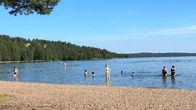 Kuva hiekkarannalta ja järvenrannasta Kalmarinrannasta Asikkalasta. Aurinko paistaa, ihmiset seisovat vedessä, uivat, lapset leikkivät.