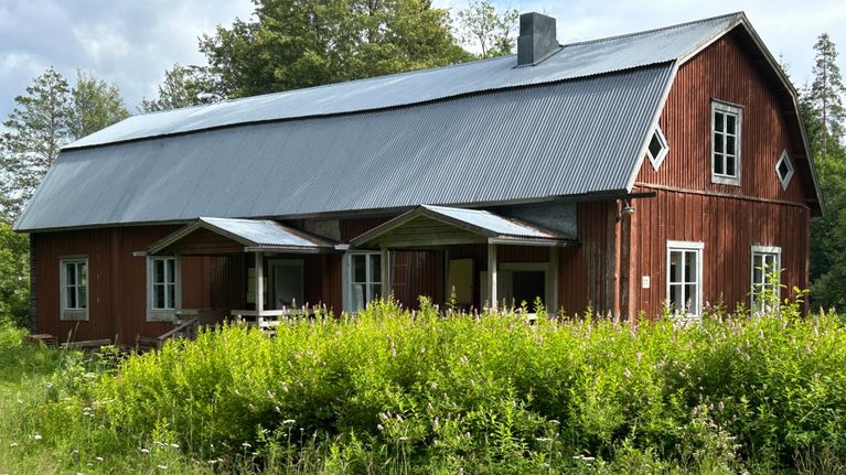 Vanha punainen maalaistalo vehreän luonnon ympäröimänä, jossa kukkivat kesäiset kedon kasvit.