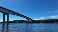 Vesistön yli kulkee korkea silta, ja taustalla näkyy vene sekä metsäinen ranta kirkkaassa auringonpaisteessa.