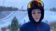 Mäkihyppyhaalariin ja kypärään pukeutunut henkilö seisoo mäkihyppyrin edustalla.