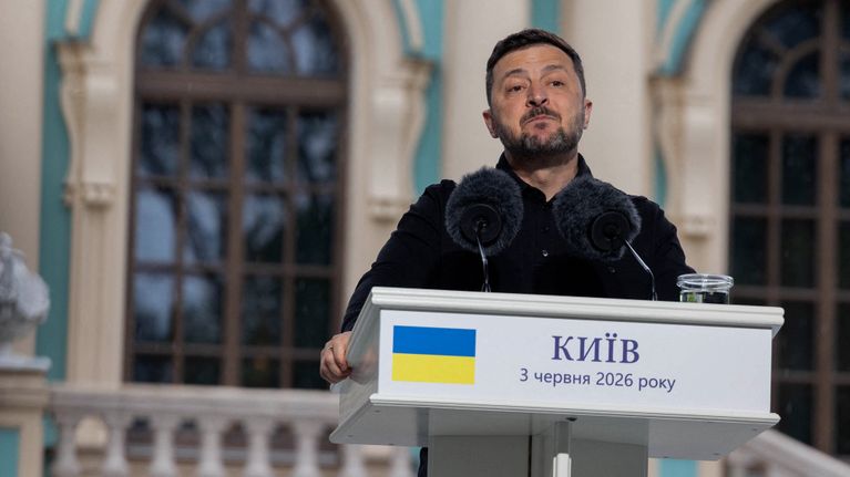 Zelenskyi seisoo puhujanpöntön takana, edessään kaksi kookasta mikrofonia, joissa on pörröiset tuulisuojukset.