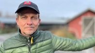 En man med keps och grön jacka står med utsträckt arm vid ett farmområde