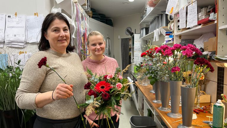 Floristit Kati Koskinen ja Riikka Jaakkola kukkakauppa Helmililjan takahuonessa. Koskisella on kimppu kädessä, jota hän parhaillaan sitoo. Maljakoissa on lisää kukkia työpöydällä.