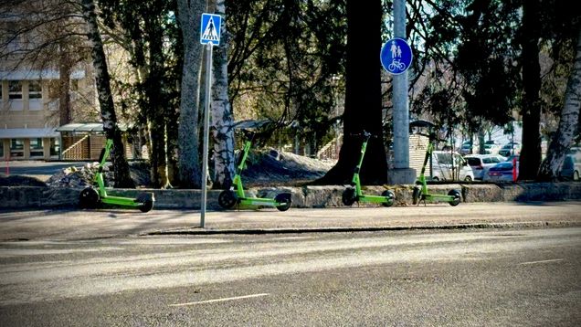 Fyra elsparkcyklar står vid en trottoar intill träd, med skyltar för övergångsställe och gång- och cykelväg.