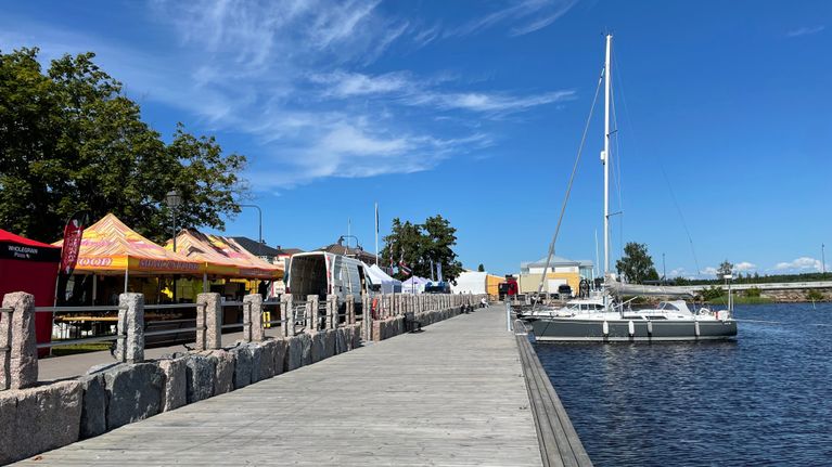 En segelbåt ligger förtöjd vid en brygga som löper längs en solig strandpromenad.
