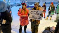 Female protesters stand holding placards in a snowy landscape, with cars and buildings in the background.
