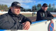 Kaksi miestä nojaa urheilukaukalon reunaan ja heidän välistään takana näkyy kaksi jalkapalloa pelaavaa lasta.