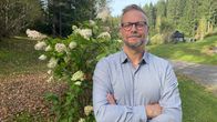 Silmälasipäinen mies seisoo kädet eteensä ristittyinä kauniissa syksyisessä maisemassa, taustalla vihreitä puita ja kukkiva syyshortensia.