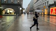 A fairly empty city street on a rainy day, with a few people walking on the sidewalk, a woman in the foreground is holding an umbrella.