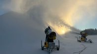 Kaksi keltaista lumitykkiä täydessä tohinassa, taustalla lumen läpi hohtava aurinko.